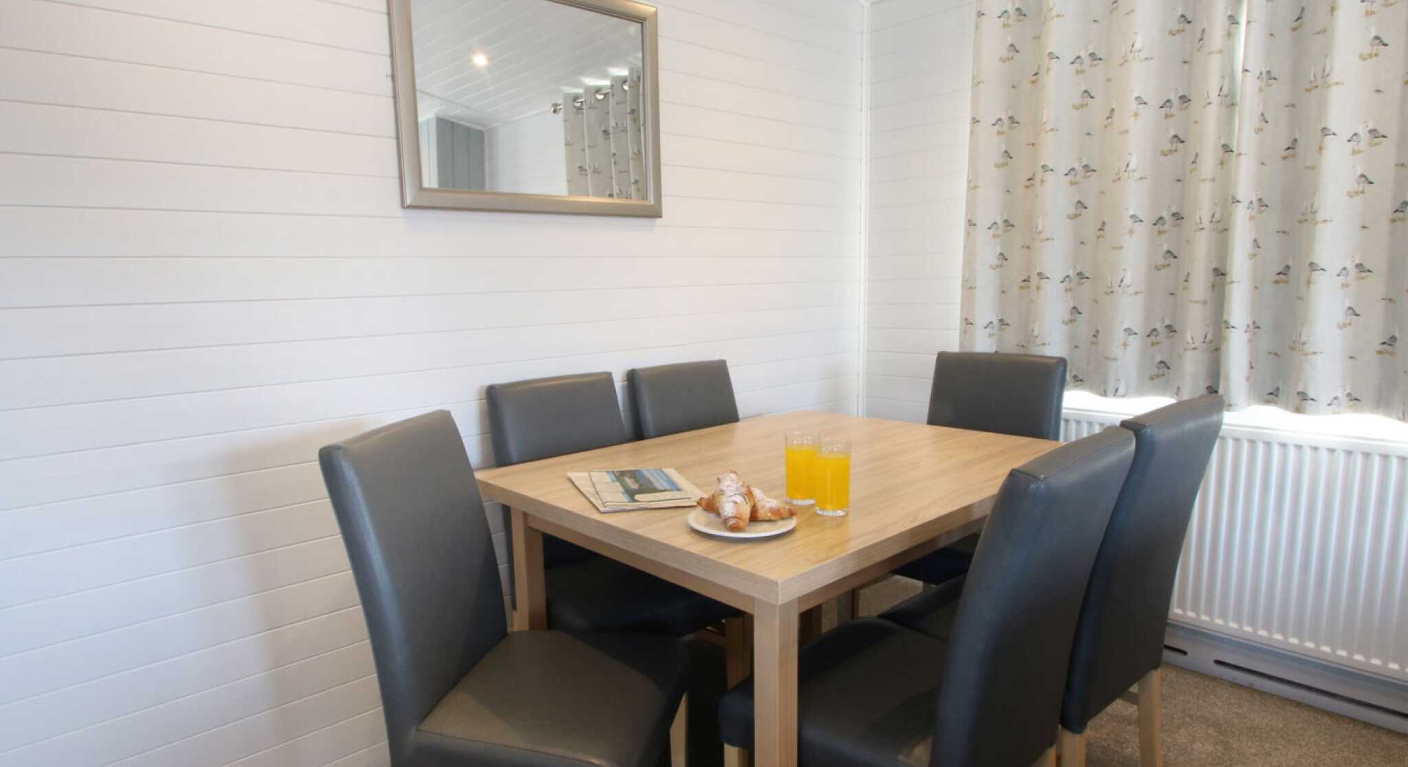 A cozy dining area with a wooden table surrounded by six black leather chairs. A plate with pastries and two glasses of orange juice sit on the table. A mirror hangs on the wall, reflecting the soft light from a nearby window, which is dressed with patterned curtains.