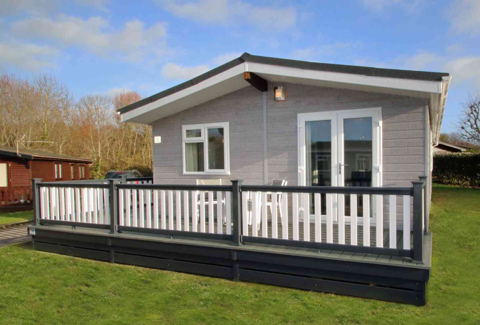 A modern cabin with a gray exterior and a large deck, surrounded by greenery. The front features large windows and double doors leading to the deck, which has white railings.