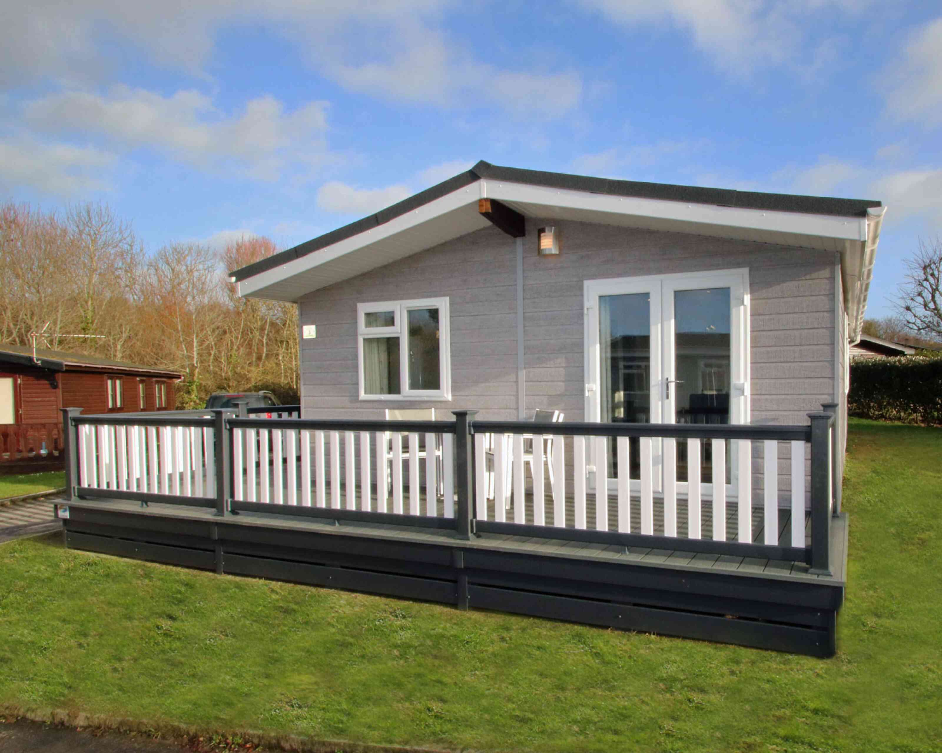 A modern cabin with a gray exterior and a large deck, surrounded by greenery. The front features large windows and double doors leading to the deck, which has white railings.