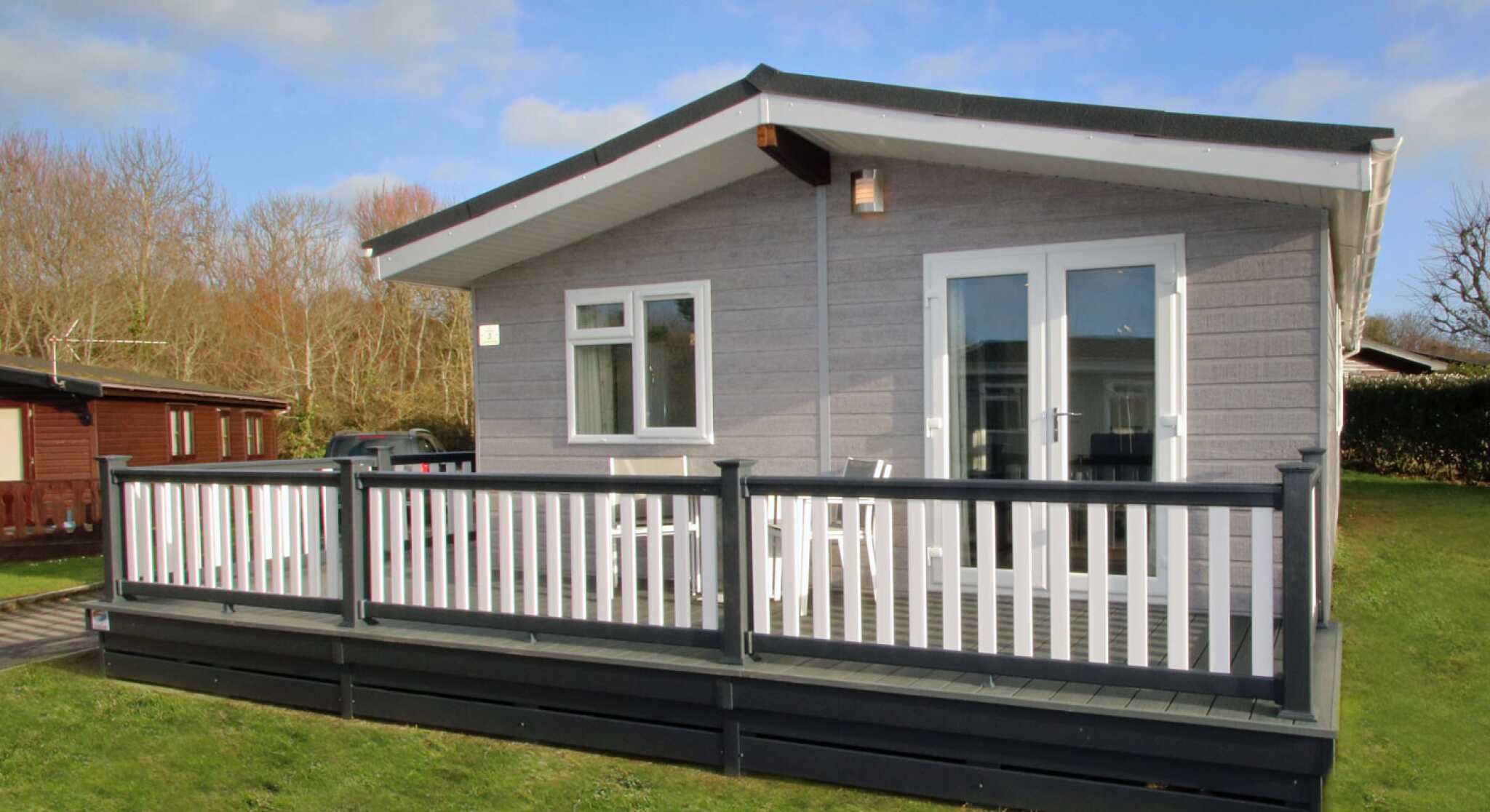A modern cabin with a gray exterior and a large deck, surrounded by greenery. The front features large windows and double doors leading to the deck, which has white railings.