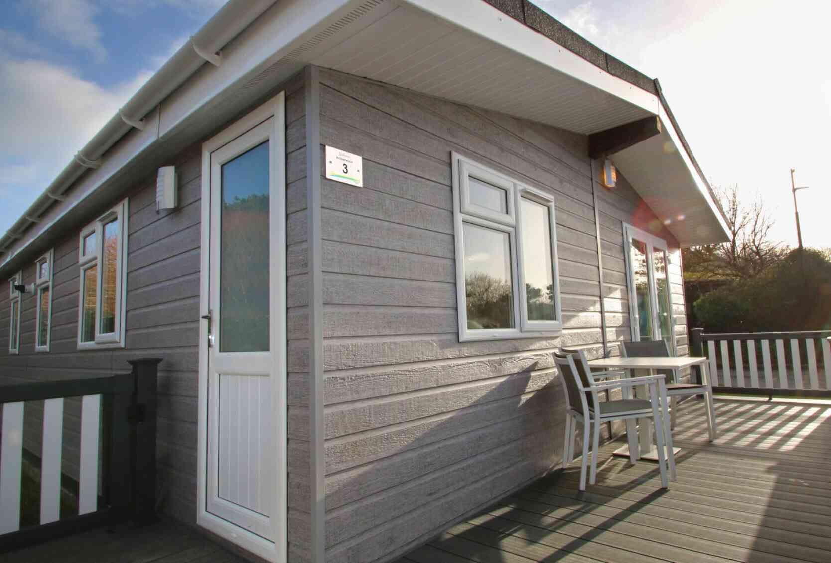 A modern, wooden cabin with horizontal siding, featuring a white door and a window, situated on a deck with a small table and chairs. The setting has clear skies and some greenery in the background.