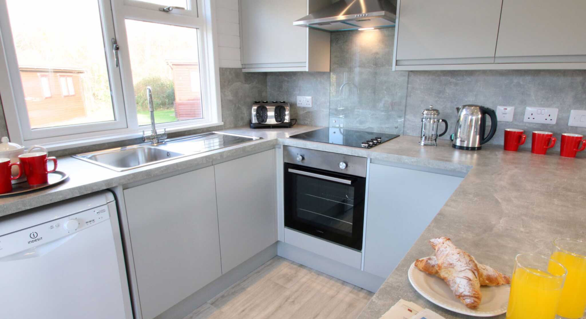 Modern kitchen with light gray cabinets and appliances. Includes a window showing a view outside, a sink, a stove, and a kettle. A plate with croissants and two glasses of orange juice are on the counter, alongside a newspaper. Red mugs are placed on the countertop.