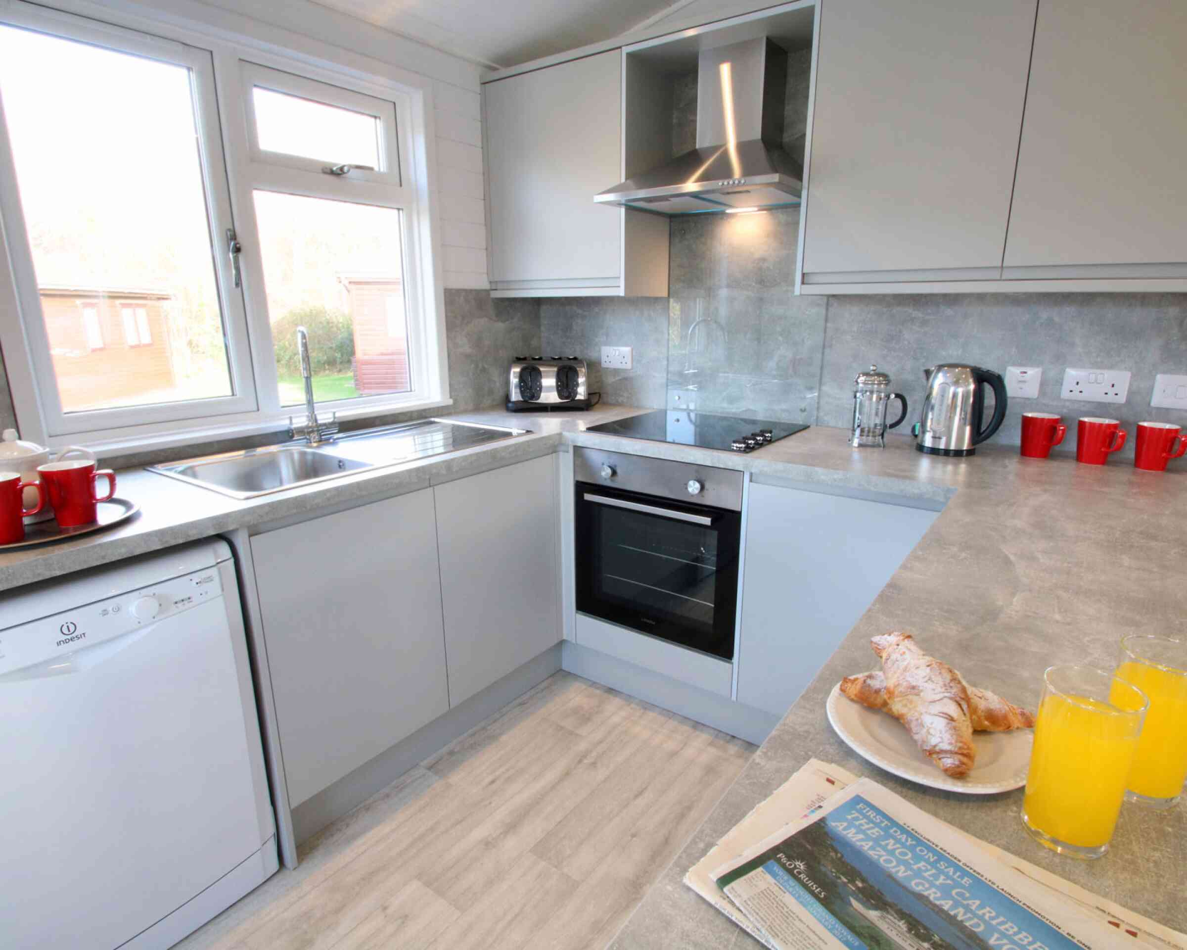 Modern kitchen with light gray cabinets and appliances. Includes a window showing a view outside, a sink, a stove, and a kettle. A plate with croissants and two glasses of orange juice are on the counter, alongside a newspaper. Red mugs are placed on the countertop.