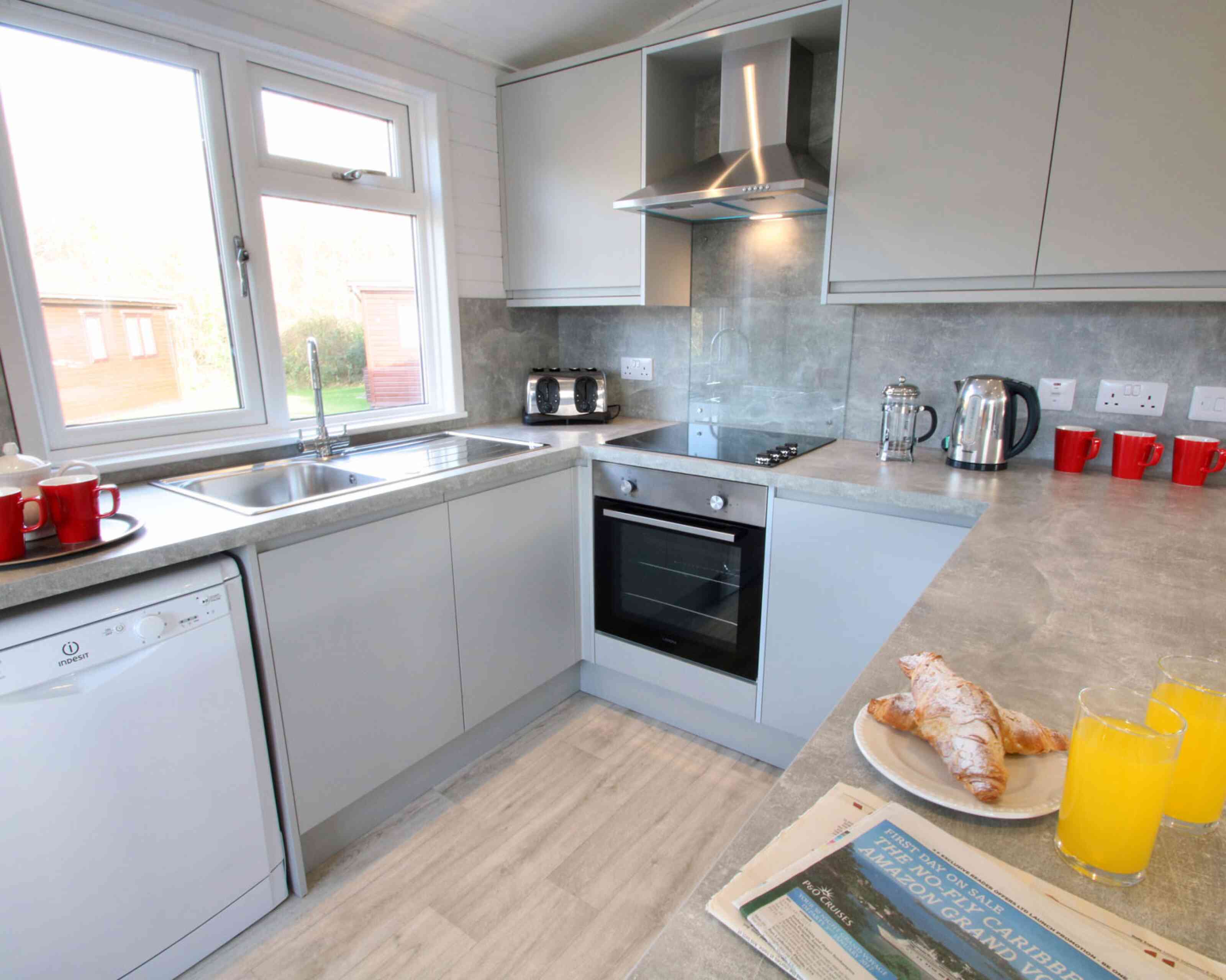 Modern kitchen with light gray cabinets and appliances. Includes a window showing a view outside, a sink, a stove, and a kettle. A plate with croissants and two glasses of orange juice are on the counter, alongside a newspaper. Red mugs are placed on the countertop.
