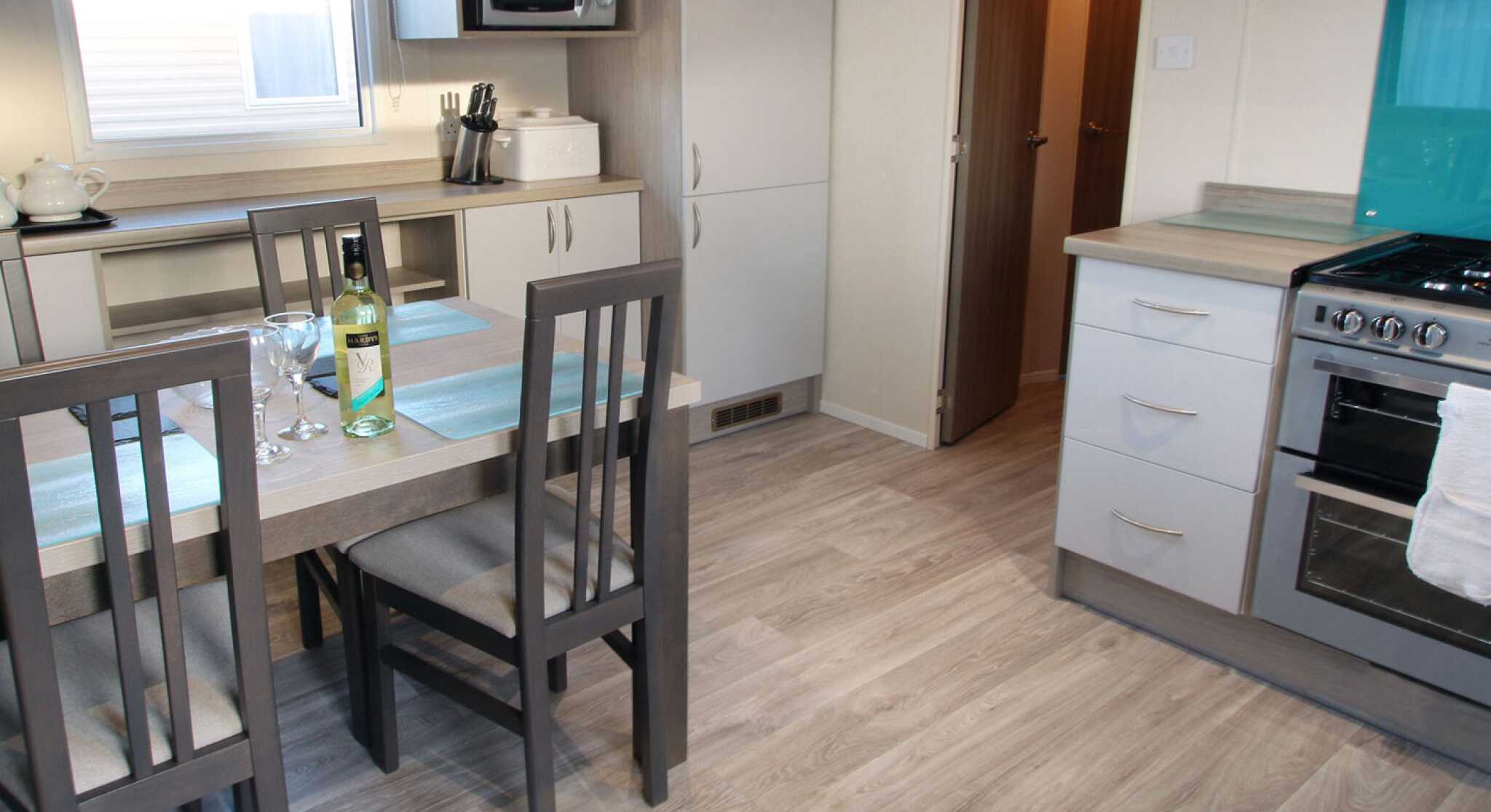 A modern kitchen features a dining table set for two with a bottle of wine and glasses. Gray cabinets provide storage, while a turquoise backsplash contrasts with the countertop. A stove and microwave are visible, along with a doorway leading to another room.