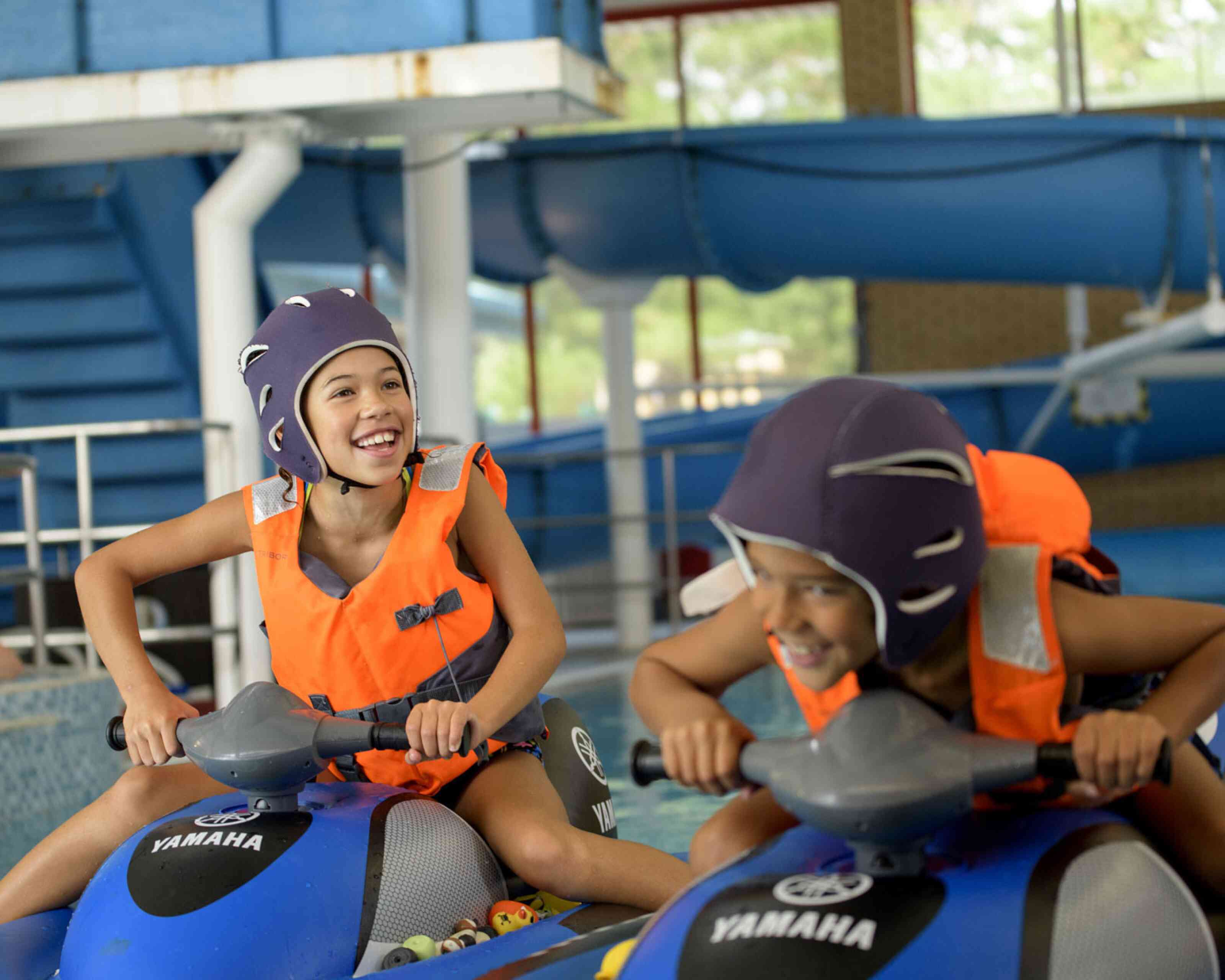 Two smiling children wearing life jackets and helmets ride jet skis indoors, surrounded by a water slide and the blue of a swimming pool. They look excited and engaged in their activity.