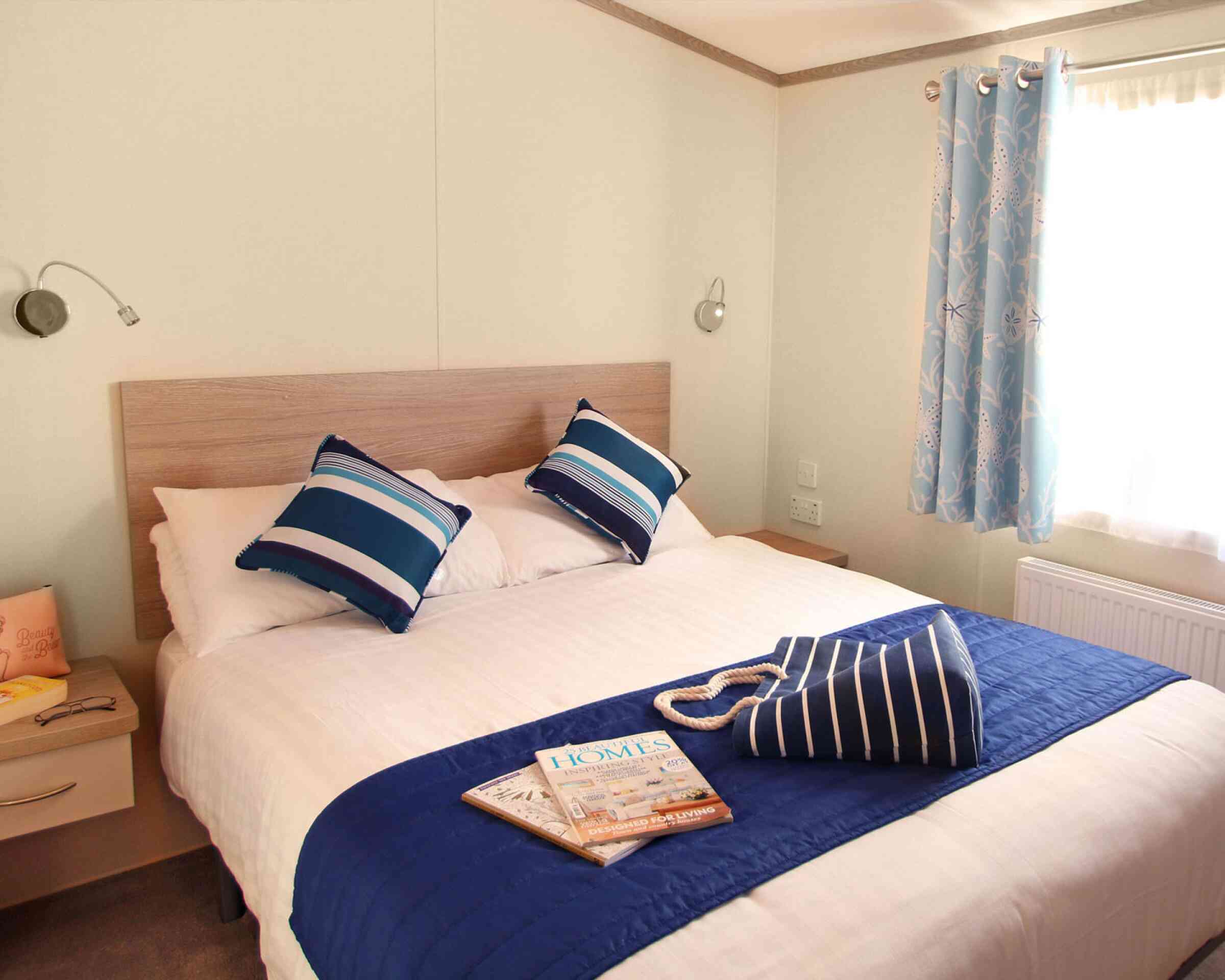 A cozy bedroom features a large bed with white linens and blue-accented pillows. A decorative blue throw is draped over the bed. A bedside table holds a magazine, and light blue curtains frame a window.