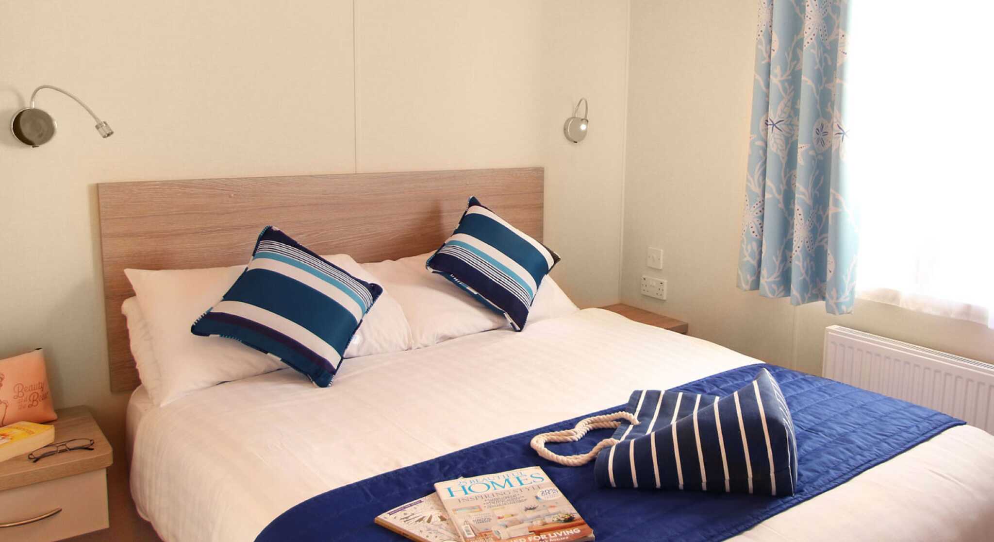 A cozy bedroom features a large bed with white linens and blue-accented pillows. A decorative blue throw is draped over the bed. A bedside table holds a magazine, and light blue curtains frame a window.