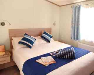 A cozy bedroom features a large bed with white linens and blue-accented pillows. A decorative blue throw is draped over the bed. A bedside table holds a magazine, and light blue curtains frame a window.