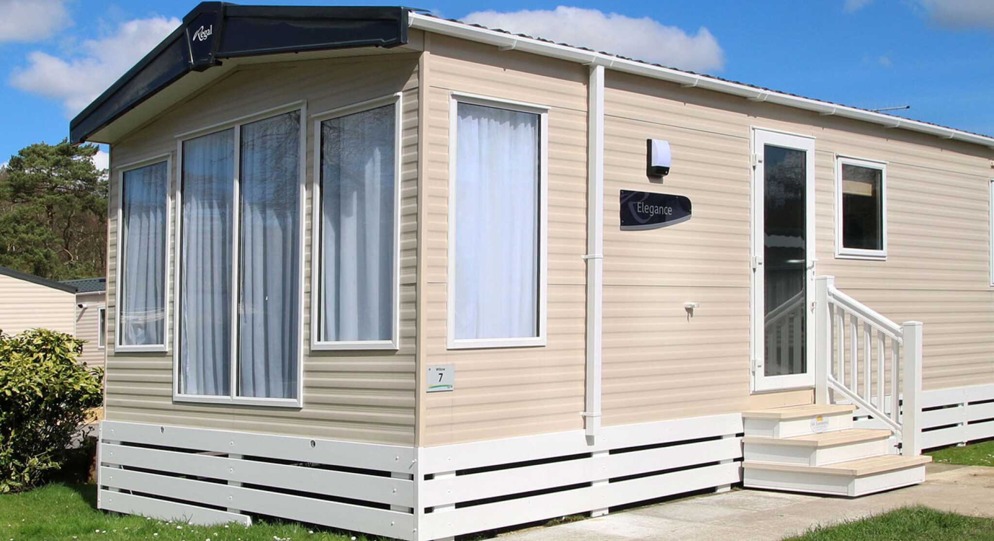 A modern beige mobile home with large windows and a white front porch. The home features a sign reading "Elegance" and is surrounded by green grass and shrubs under a sunny blue sky with fluffy clouds.