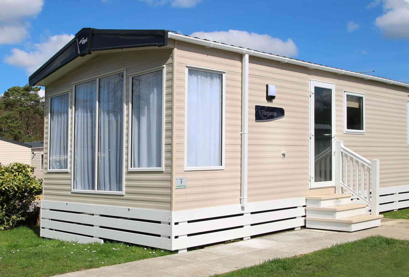 A modern beige mobile home with large windows and a white front porch. The home features a sign reading "Elegance" and is surrounded by green grass and shrubs under a sunny blue sky with fluffy clouds.