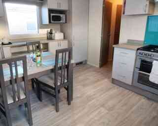 A modern kitchen featuring a dining table with four chairs, a microwave, and a stove. Light wood flooring complements the gray cabinets and turquoise backsplash. A door leads to another room, while a window provides natural light.