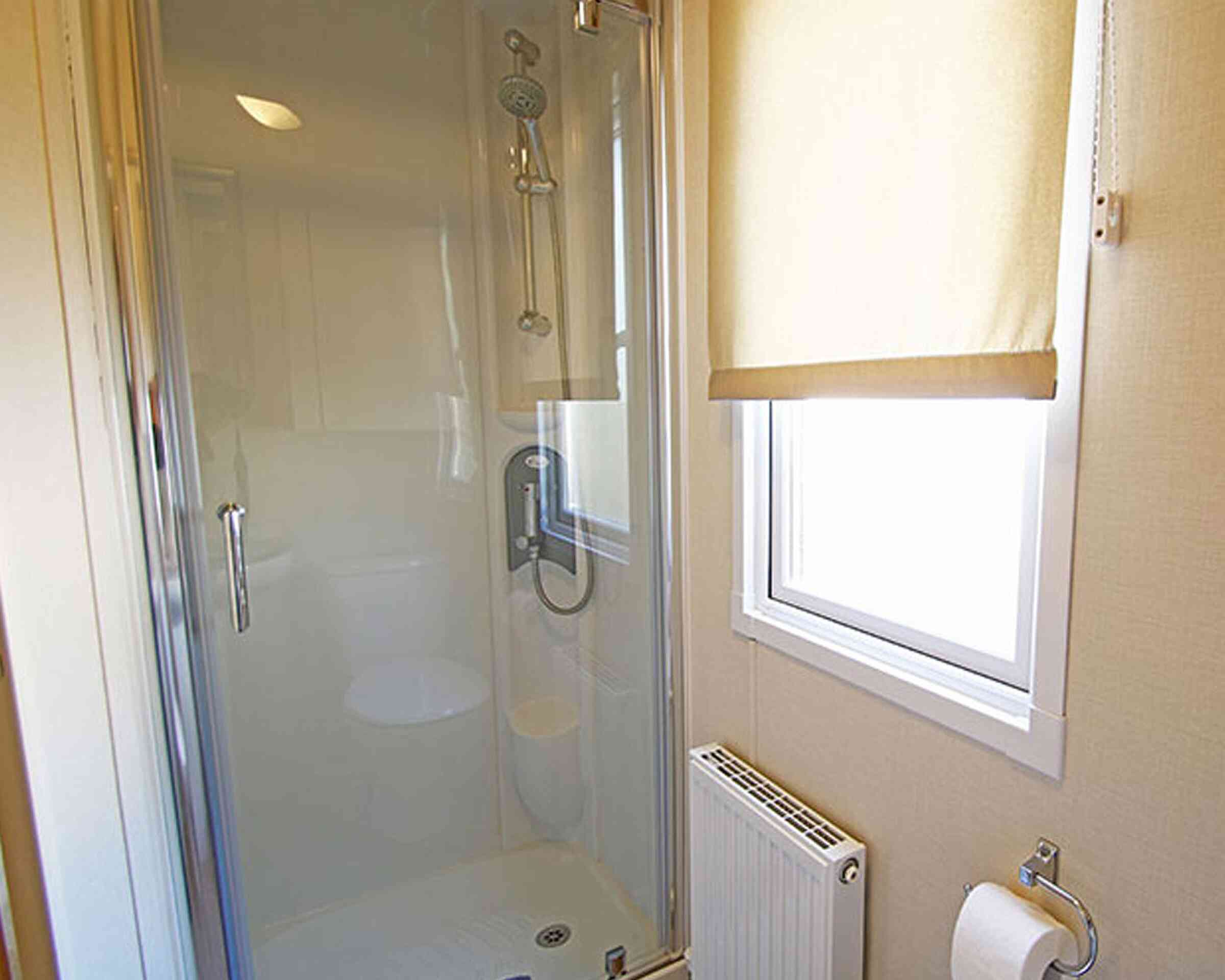 A modern bathroom shower stall featuring clear glass doors, a showerhead, and a sleek faucet. A window with a beige roller shade allows natural light, and a towel rack is mounted nearby. A radiator is visible, providing warmth.