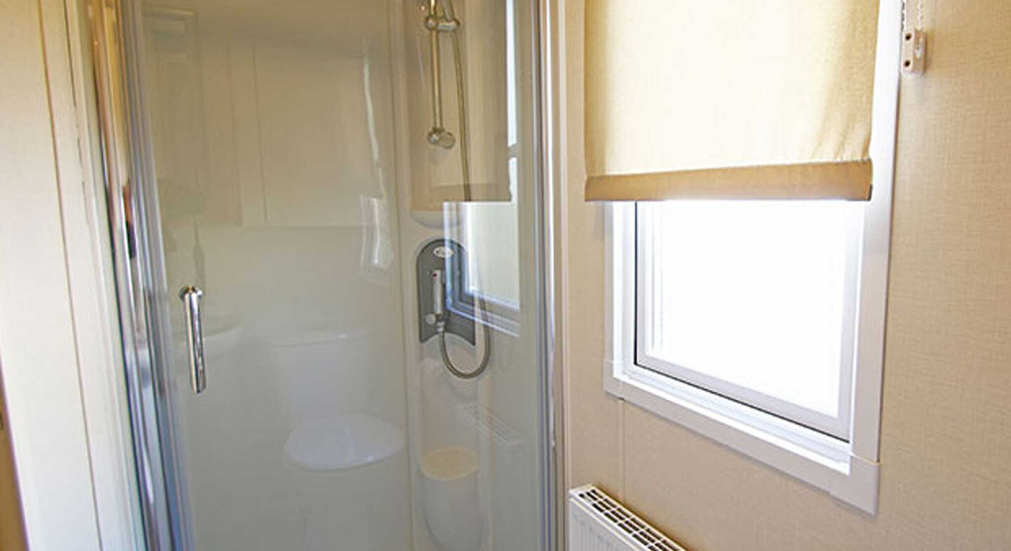 A modern bathroom shower stall featuring clear glass doors, a showerhead, and a sleek faucet. A window with a beige roller shade allows natural light, and a towel rack is mounted nearby. A radiator is visible, providing warmth.
