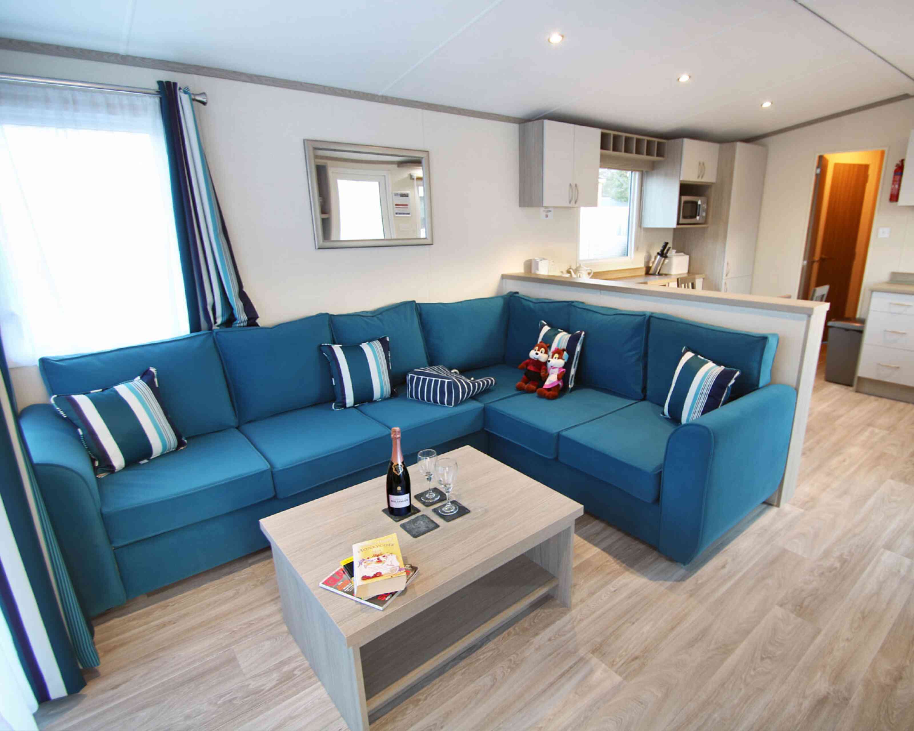 A modern living room features a large blue sectional sofa with decorative pillows, a light wooden coffee table with drinks and books, and a bright window with curtains. The kitchen area is visible in the background.