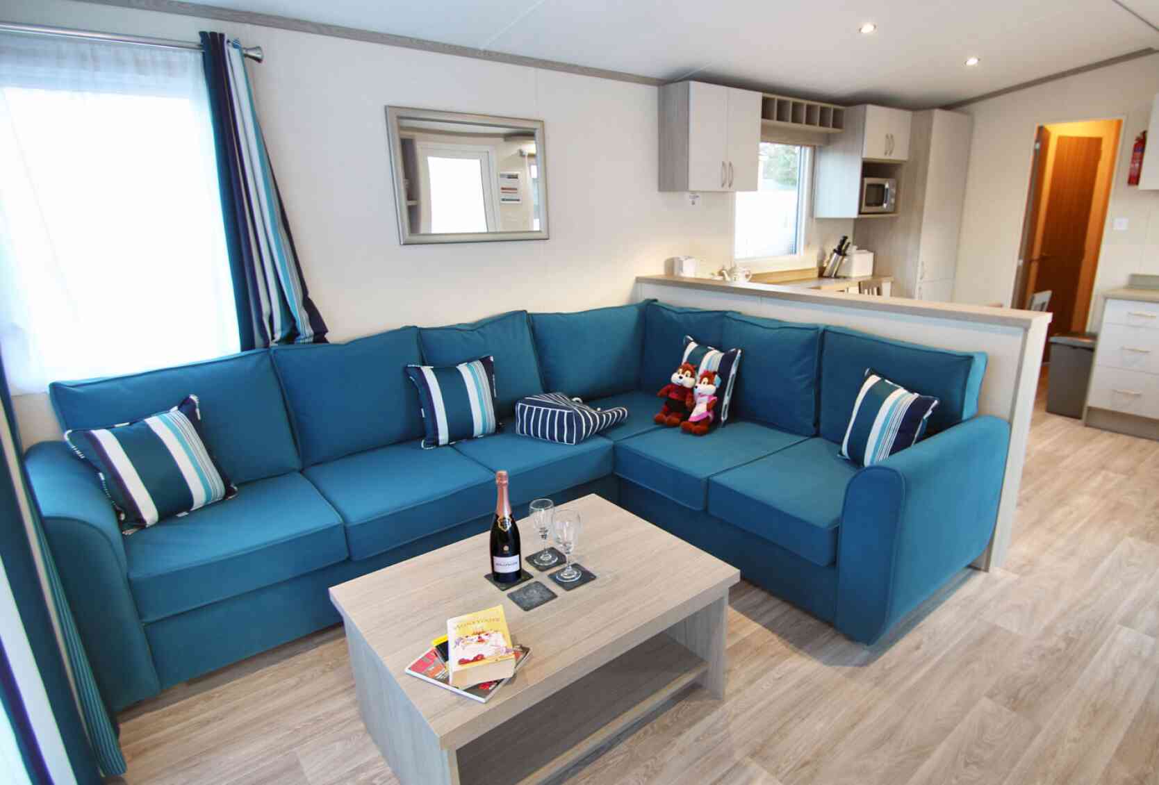 A modern living room features a large blue sectional sofa with decorative pillows, a light wooden coffee table with drinks and books, and a bright window with curtains. The kitchen area is visible in the background.