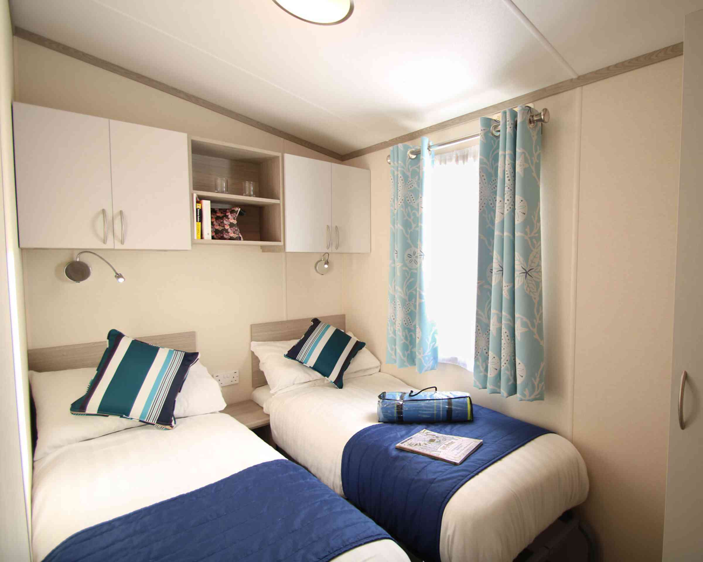 A cozy bedroom featuring two single beds with white bedding and navy blue throw blankets. There are blue and white patterned curtains at the window, and shelving above the beds with books and decorative items. A magazine is placed on one of the beds.