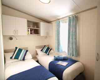 A cozy bedroom featuring two single beds with white bedding and navy blue throw blankets. There are blue and white patterned curtains at the window, and shelving above the beds with books and decorative items. A magazine is placed on one of the beds.