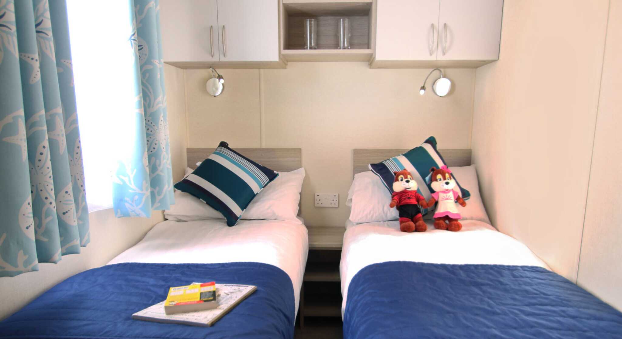 A cozy twin bedroom with two single beds, each adorned with striped blue and white pillows. A blue bedspread covers the beds, and a small table between them features a book and a decorative item. Plush toys sit on each bed. Light blue curtains with a bird pattern frame a window, providing natural light. Shelves above the beds hold decorative items.