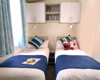 A cozy twin bedroom with two single beds, each adorned with striped blue and white pillows. A blue bedspread covers the beds, and a small table between them features a book and a decorative item. Plush toys sit on each bed. Light blue curtains with a bird pattern frame a window, providing natural light. Shelves above the beds hold decorative items.