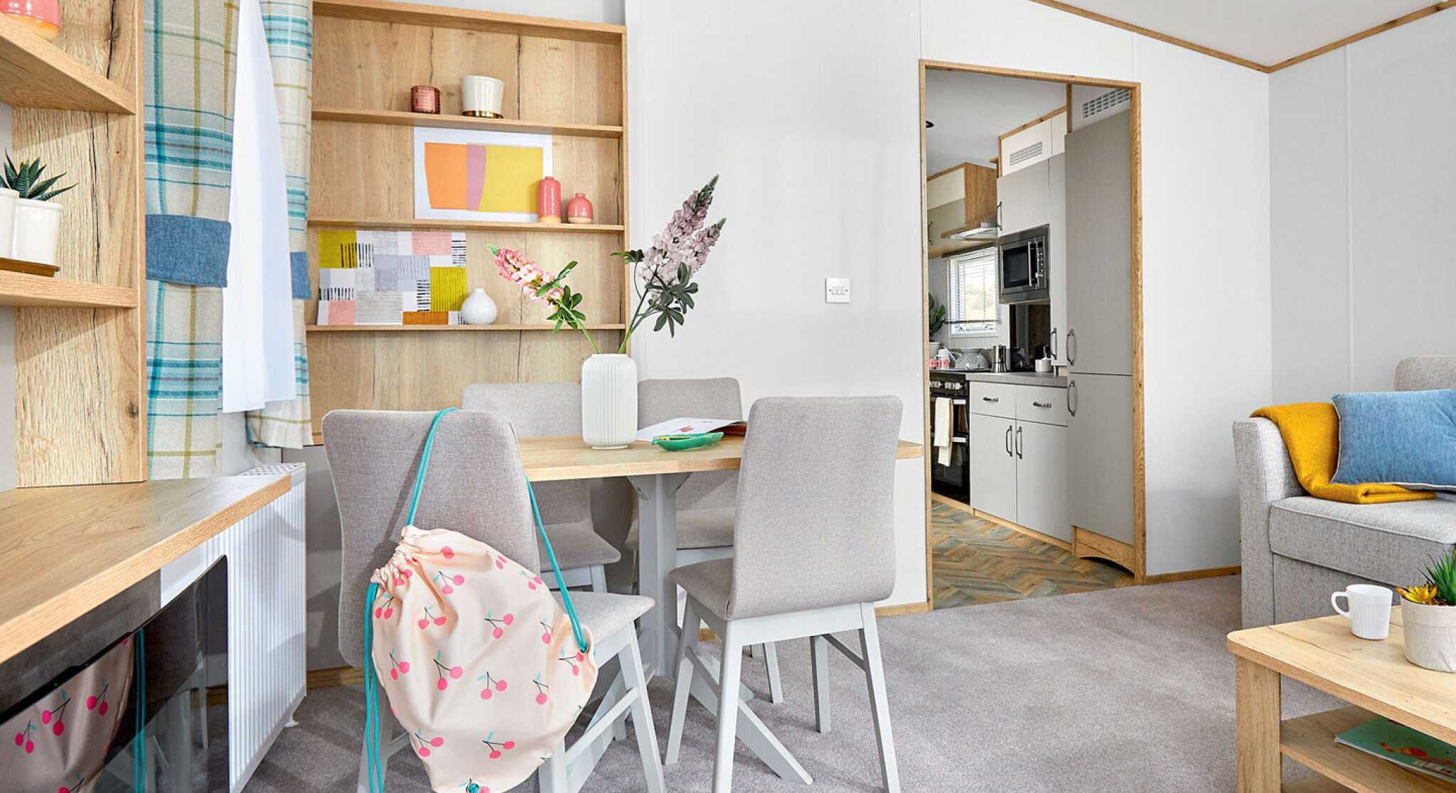 A cozy living space featuring a light wood dining table with four chairs, a colorful shelf with decorative items, and a kitchen area visible in the background. Soft gray carpeting complements the decor, which includes a blue and yellow throw on a nearby sofa.