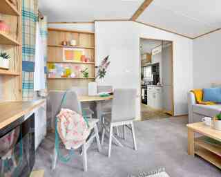 A cozy living space featuring a light wood dining table with four chairs, a colorful shelf with decorative items, and a kitchen area visible in the background. Soft gray carpeting complements the decor, which includes a blue and yellow throw on a nearby sofa.