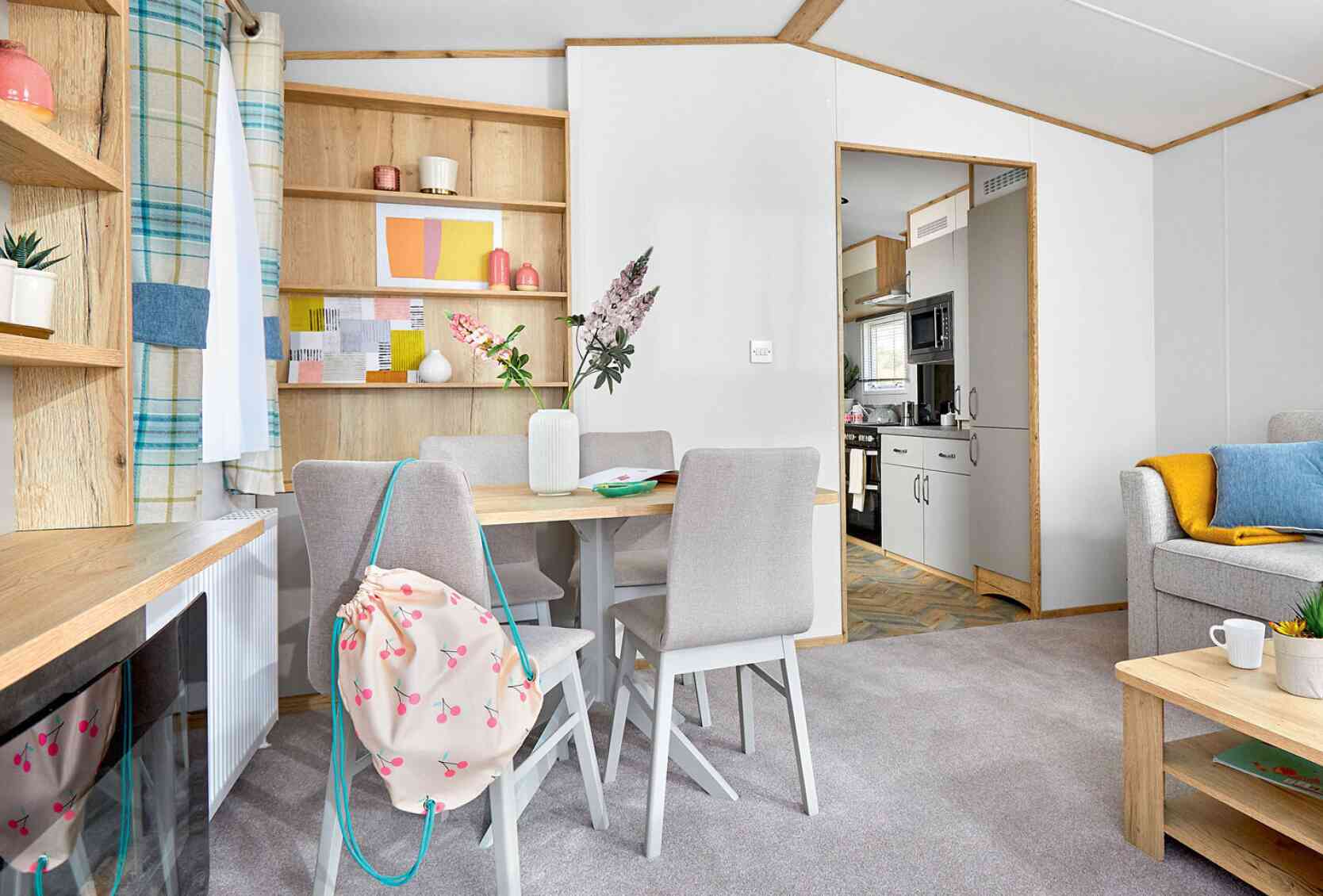 A cozy living space featuring a light wood dining table with four chairs, a colorful shelf with decorative items, and a kitchen area visible in the background. Soft gray carpeting complements the decor, which includes a blue and yellow throw on a nearby sofa.