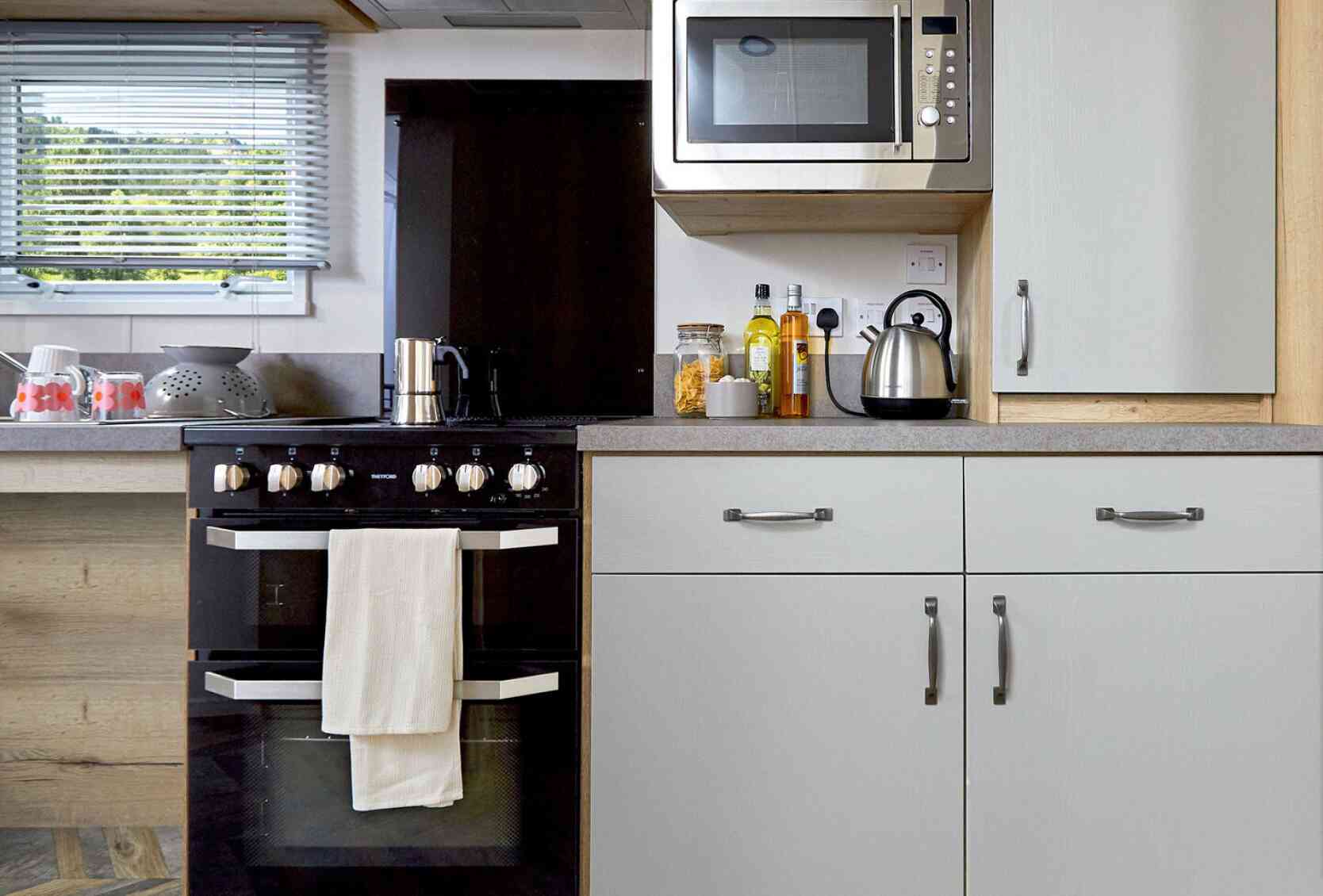 Modern kitchen featuring a black oven with a white towel, a stainless steel microwave mounted above, and a kettle on the counter. Cabinets in light grey and wooden accents complement the design. Ingredients and utensils are neatly arranged on the counter.
