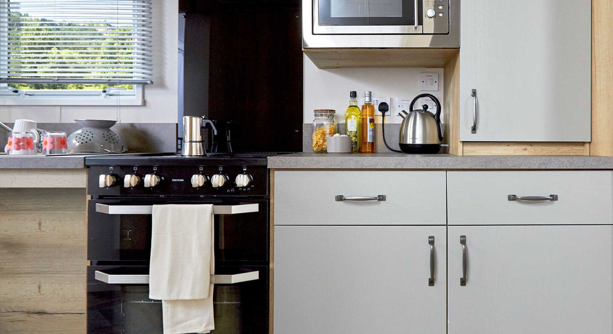 Modern kitchen featuring a black oven with a white towel, a stainless steel microwave mounted above, and a kettle on the counter. Cabinets in light grey and wooden accents complement the design. Ingredients and utensils are neatly arranged on the counter.