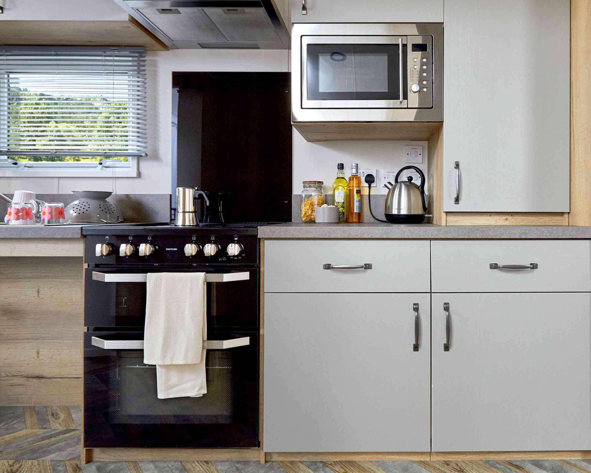Modern kitchen featuring a black oven with a white towel, a stainless steel microwave mounted above, and a kettle on the counter. Cabinets in light grey and wooden accents complement the design. Ingredients and utensils are neatly arranged on the counter.