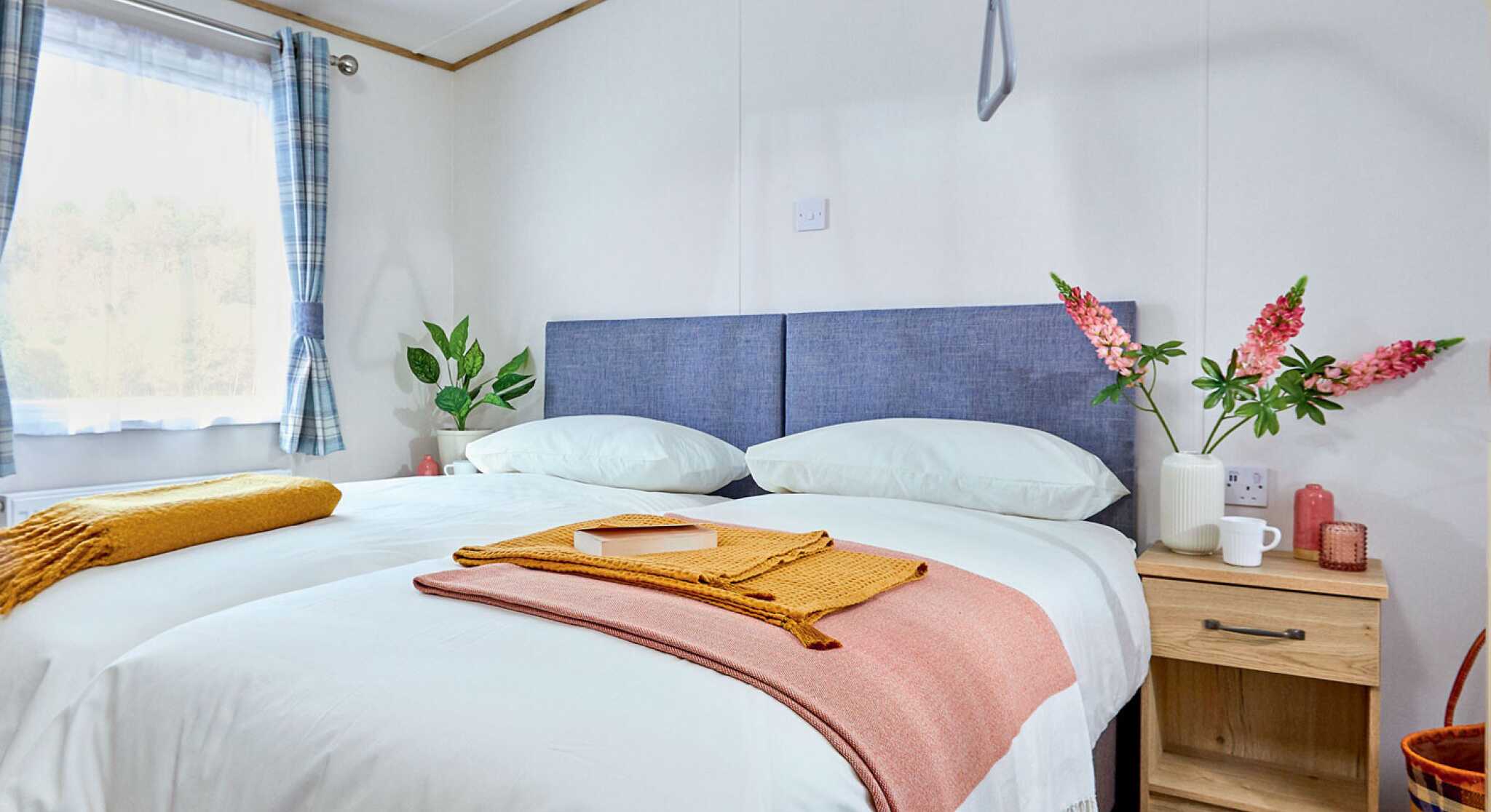 A cozy bedroom featuring a double bed with light bedding, accented by a coral throw and a gold blanket. A bedside table holds a mug and a small orange candle. Two floral arrangements add color, and a large window lets in natural light, with checkered curtains.