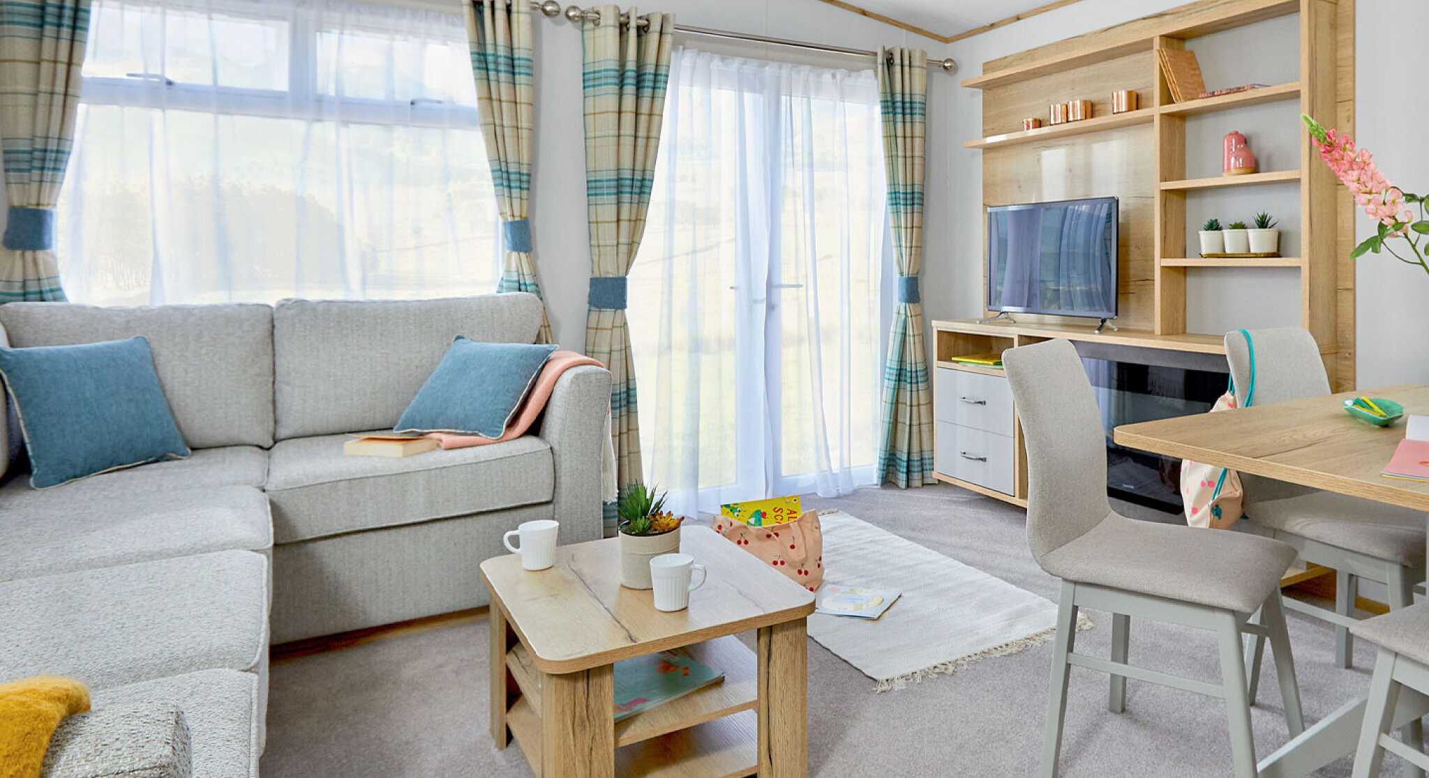 A cozy living area furnished with a light gray sectional sofa, wooden coffee table, and a small dining table with two chairs. A TV is on a wooden unit, and the room features large windows with checkered curtains, allowing natural light to brighten the space. A small plant and decorative items are on the shelves.