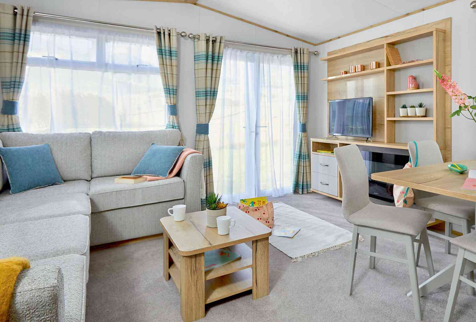 A cozy living area furnished with a light gray sectional sofa, wooden coffee table, and a small dining table with two chairs. A TV is on a wooden unit, and the room features large windows with checkered curtains, allowing natural light to brighten the space. A small plant and decorative items are on the shelves.