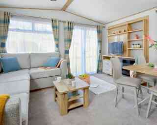 A cozy living area furnished with a light gray sectional sofa, wooden coffee table, and a small dining table with two chairs. A TV is on a wooden unit, and the room features large windows with checkered curtains, allowing natural light to brighten the space. A small plant and decorative items are on the shelves.