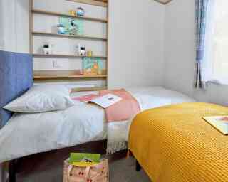 A cozy bedroom featuring two beds, one with a white blanket and a coral throw, and the other covered with a bright yellow knitted blanket. A wooden shelf holds decorative items and books, while a few magazines are spread on the bed. A window provides natural light, illuminating the space.