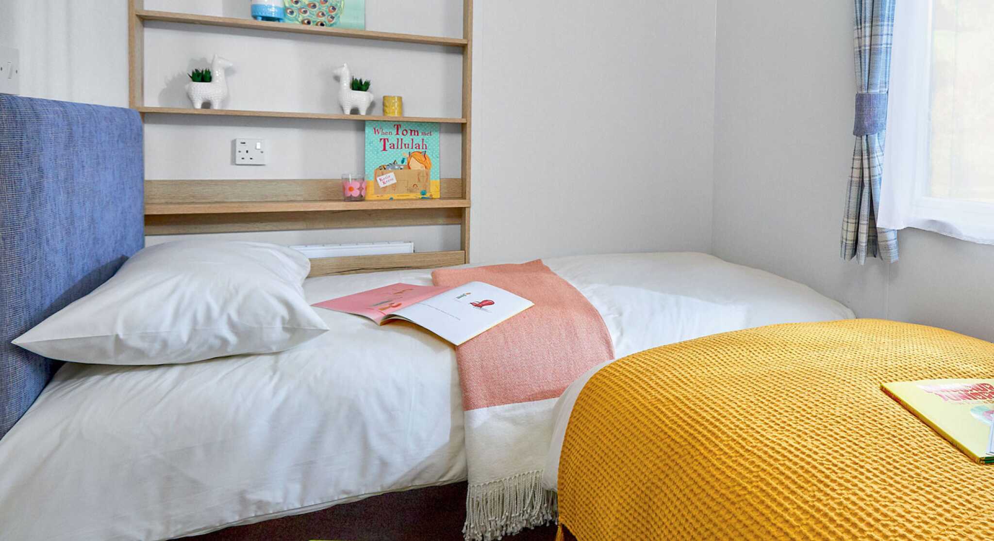 A cozy bedroom featuring two beds, one with a white blanket and a coral throw, and the other covered with a bright yellow knitted blanket. A wooden shelf holds decorative items and books, while a few magazines are spread on the bed. A window provides natural light, illuminating the space.