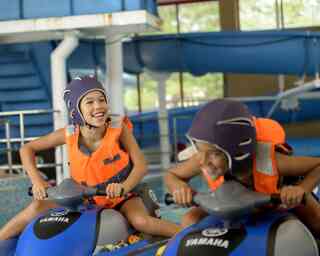 Two children wearing purple helmets and orange life vests are having fun riding water scooters in an indoor aquatic center. They are smiling and appear to be racing each other.