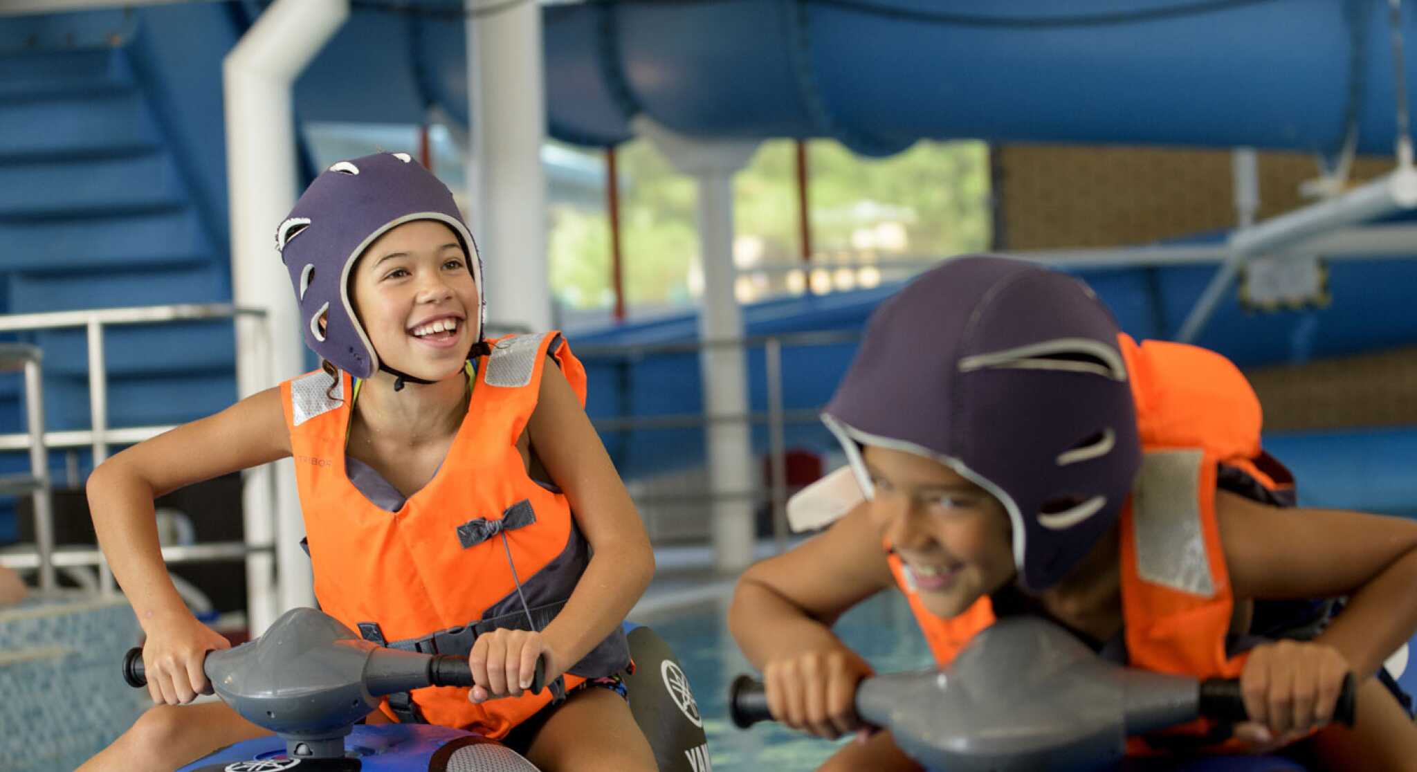Two children wearing purple helmets and orange life vests are having fun riding water scooters in an indoor aquatic center. They are smiling and appear to be racing each other.