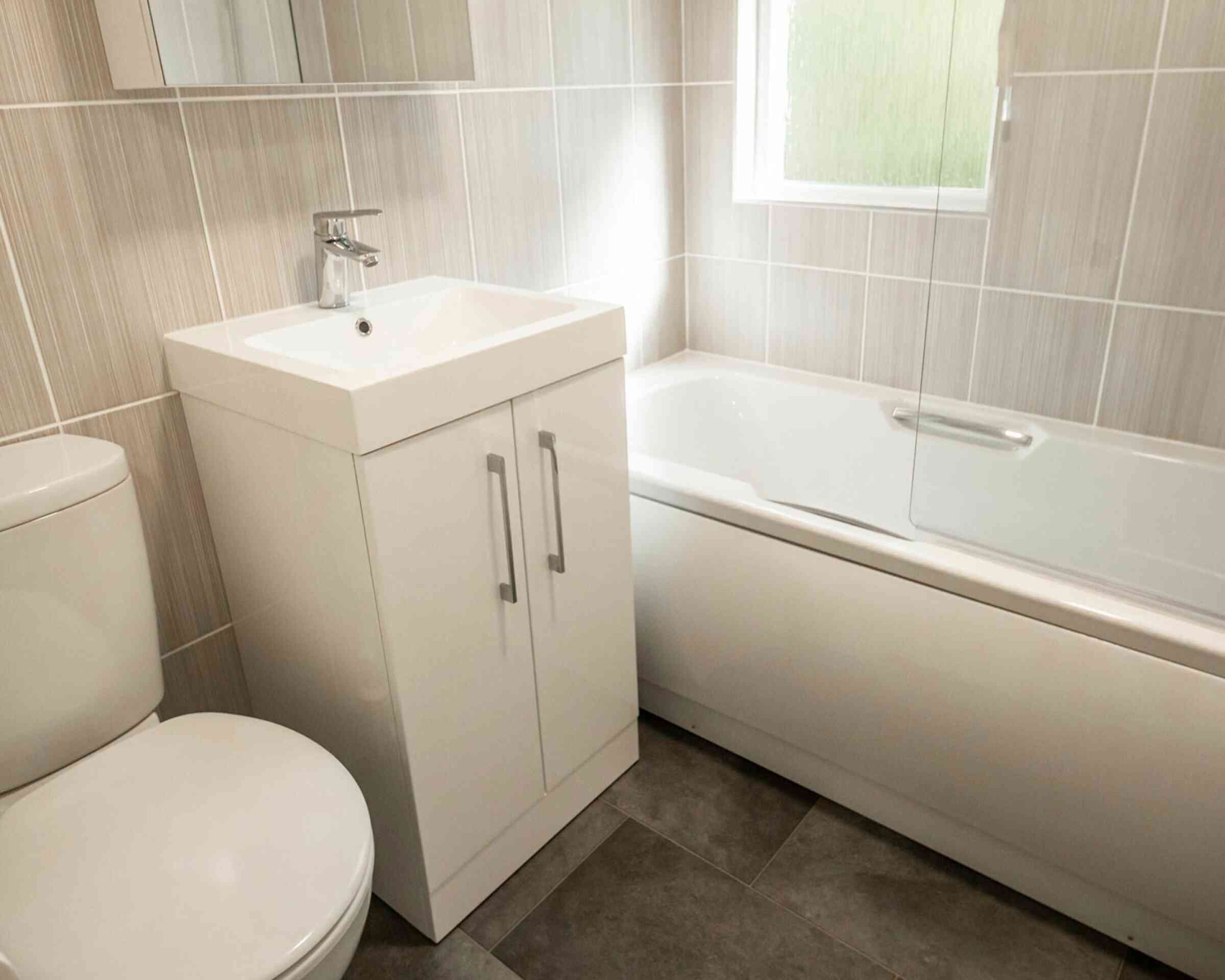 A modern bathroom featuring a white toilet, a sleek vanity with a square sink, and a bathtub with a glass screen. The walls and floor have neutral-colored tiles, with natural light coming through a window.