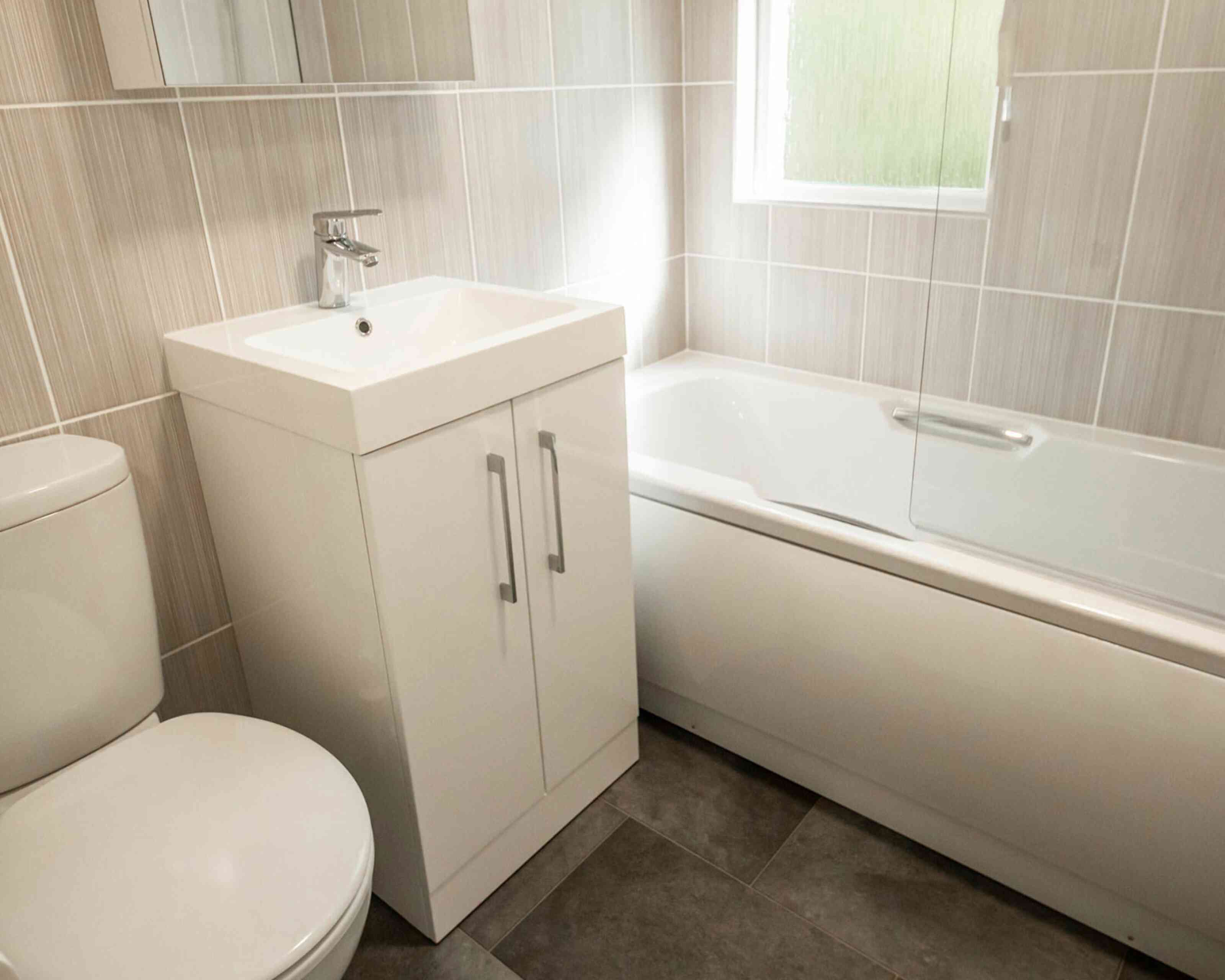 A modern bathroom featuring a white toilet, a sleek vanity with a square sink, and a bathtub with a glass screen. The walls and floor have neutral-colored tiles, with natural light coming through a window.