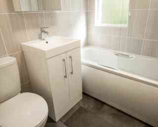 A modern bathroom featuring a white toilet, a sleek vanity with a square sink, and a bathtub with a glass screen. The walls and floor have neutral-colored tiles, with natural light coming through a window.