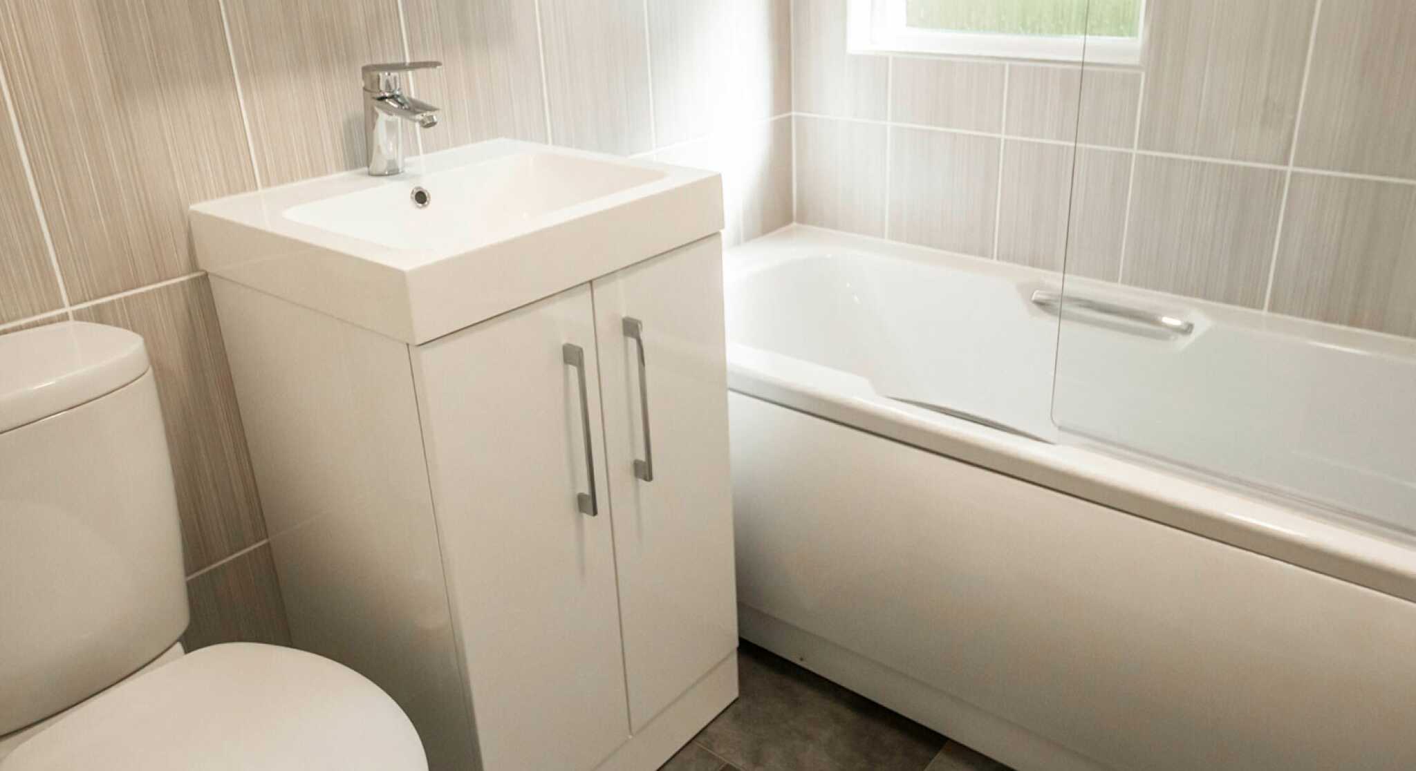 A modern bathroom featuring a white toilet, a sleek vanity with a square sink, and a bathtub with a glass screen. The walls and floor have neutral-colored tiles, with natural light coming through a window.