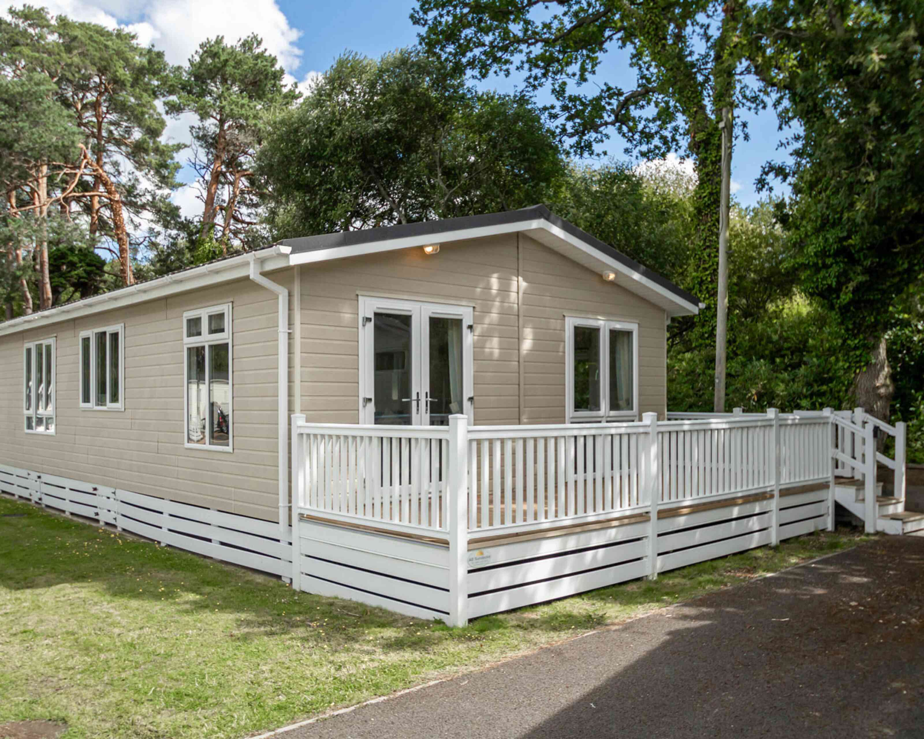 A modern single-story cabin with a light-colored wooden exterior and a spacious front deck. The cabin is surrounded by greenery and tall trees.