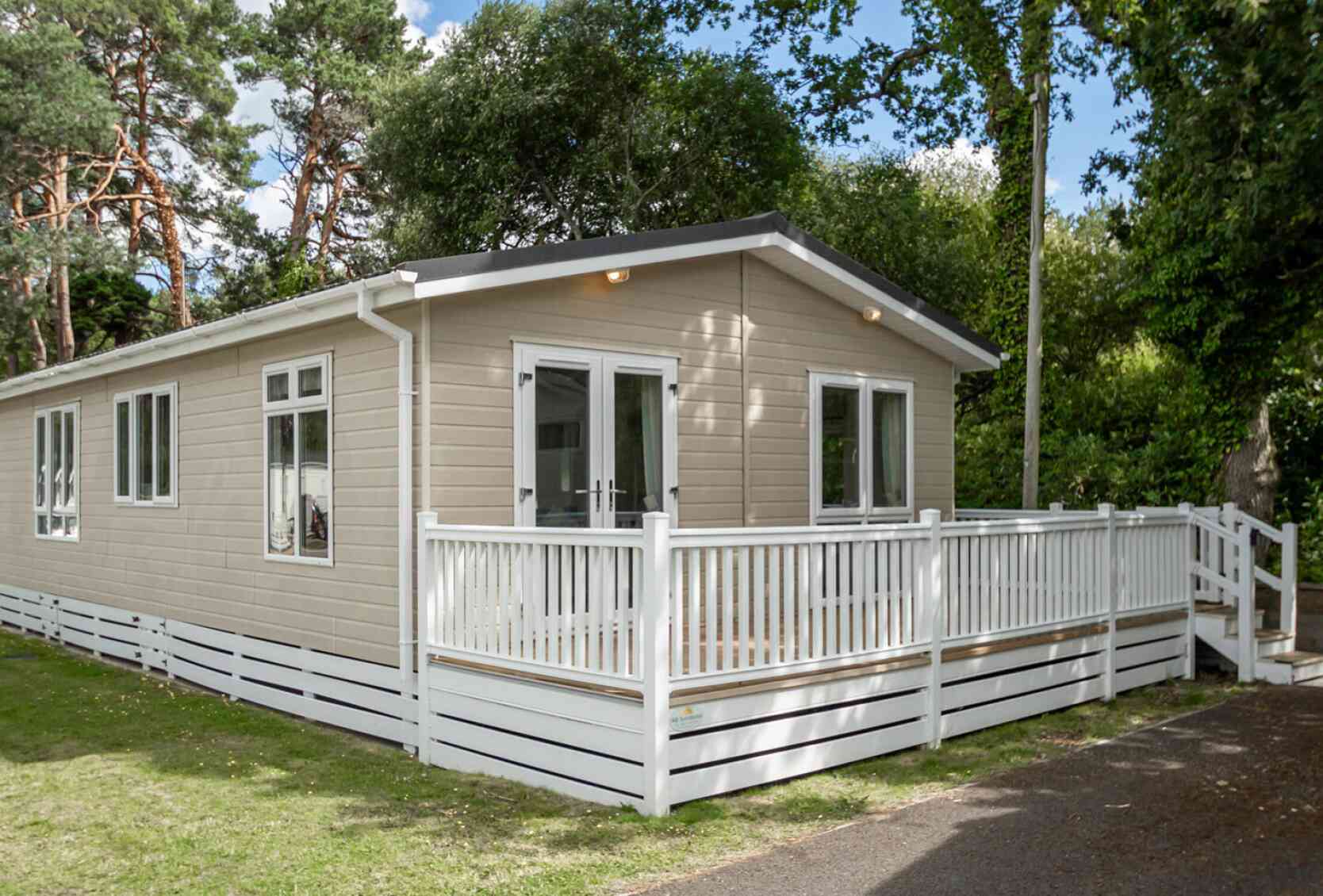 A modern single-story cabin with a light-colored wooden exterior and a spacious front deck. The cabin is surrounded by greenery and tall trees.