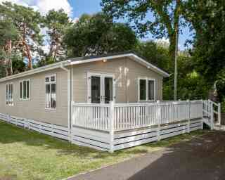 A modern single-story cabin with a light-colored wooden exterior and a spacious front deck. The cabin is surrounded by greenery and tall trees.