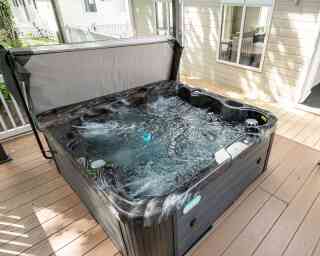 A hot tub with bubbling water sits on a wooden deck, surrounded by light-colored walls of nearby buildings. Natural light illuminates the scene.