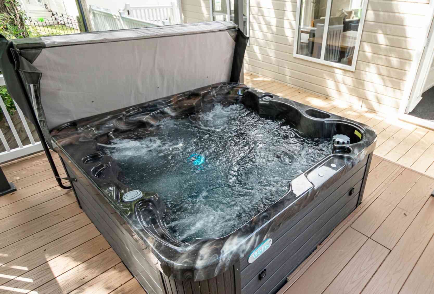A hot tub with bubbling water sits on a wooden deck, surrounded by light-colored walls of nearby buildings. Natural light illuminates the scene.
