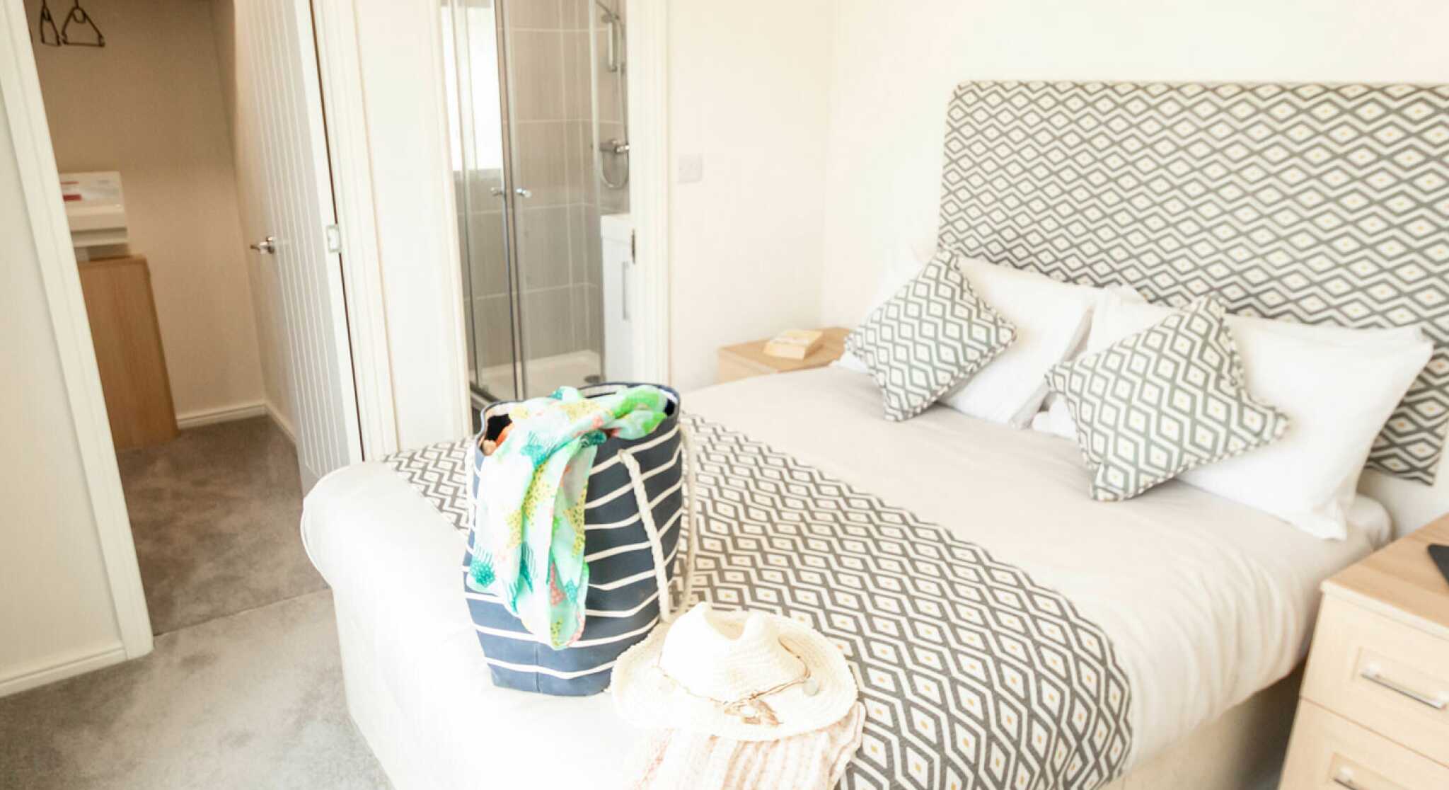 A stylish bedroom featuring a king-sized bed with patterned bedding and two decorative pillows. A packed bag and a sun hat rest on the bed. In the background, there is an en-suite bathroom with a glass shower and closet space visible. Soft carpeting adds to the cozy atmosphere.
