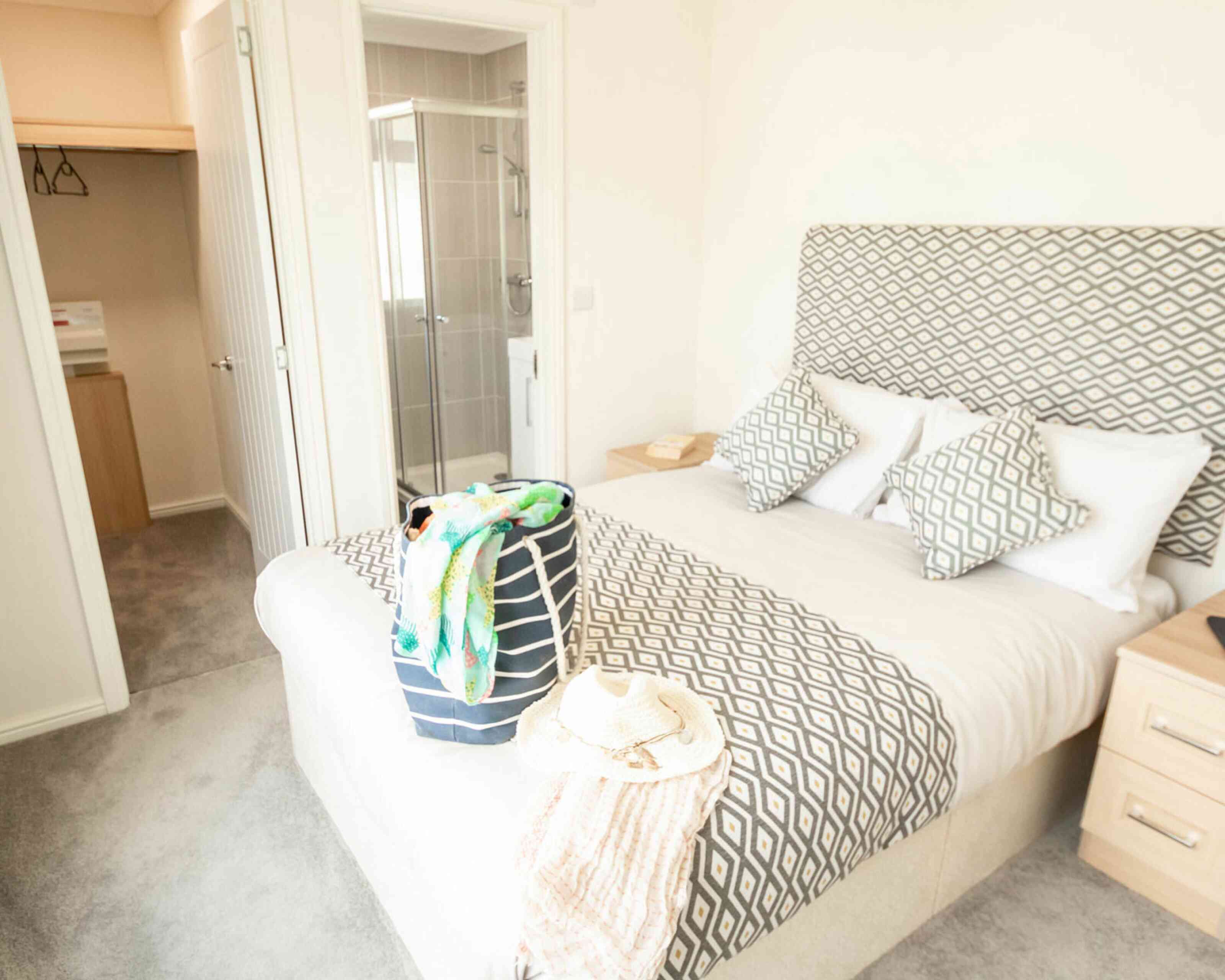 A stylish bedroom featuring a king-sized bed with patterned bedding and two decorative pillows. A packed bag and a sun hat rest on the bed. In the background, there is an en-suite bathroom with a glass shower and closet space visible. Soft carpeting adds to the cozy atmosphere.