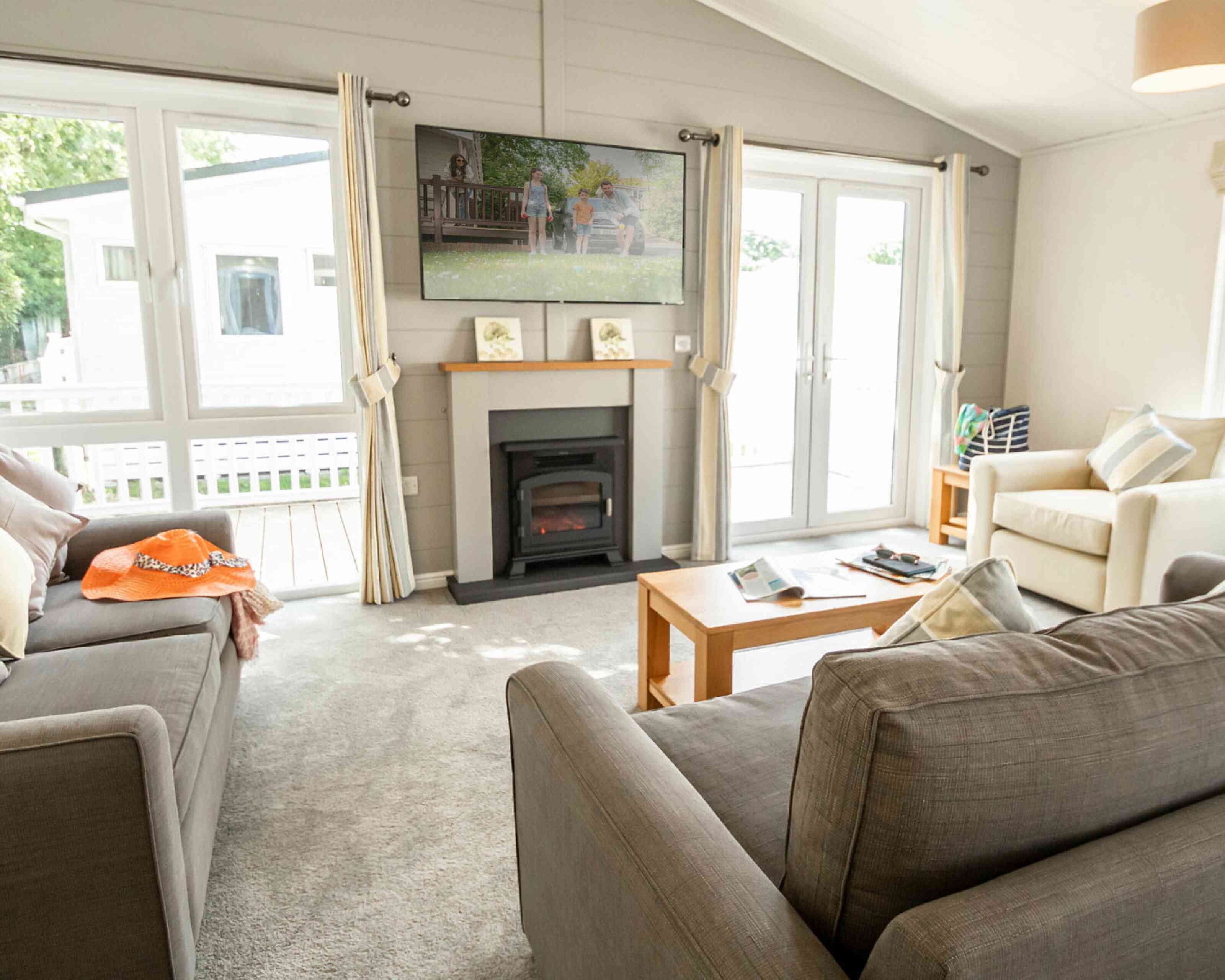 A cozy living room featuring two sofas, a coffee table, and a television mounted on the wall. Light streams in through large windows, with a view of a deck outside. Soft, neutral colors and decorative pillows create a warm atmosphere.